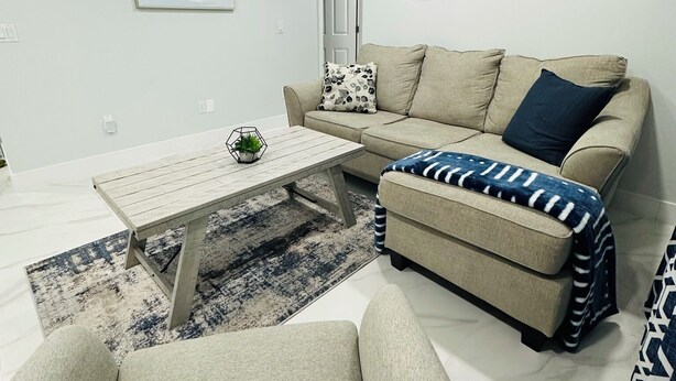 A modern living room with a sofa, ottoman, coffee table, and a rug. - Home Away From Home! (Vero Beach)