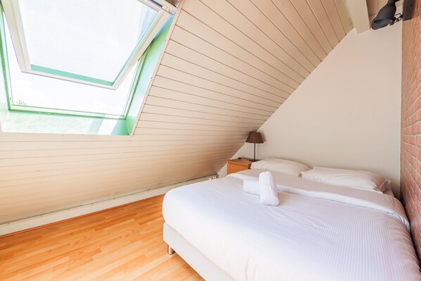 Ein Schlafzimmer mit Schrägdecke, einem Fenster, einem Bett mit weißer Bettwäsche und einem Nachttisch mit Lampe.