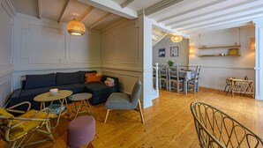 Una sala acogedora con un sofá de color oscuro, una mesita redonda y un reposapiés morado. Hay un comedor con una mesa y sillas de madera, y una cocina con una lámpara colgante.