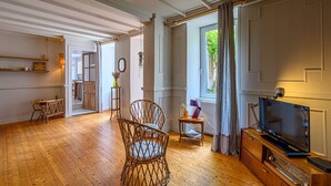 Un salón amplio con piso de madera, un televisor y un aparador de madera. Hay una puerta que da a otra habitación y una ventana con cortinas.