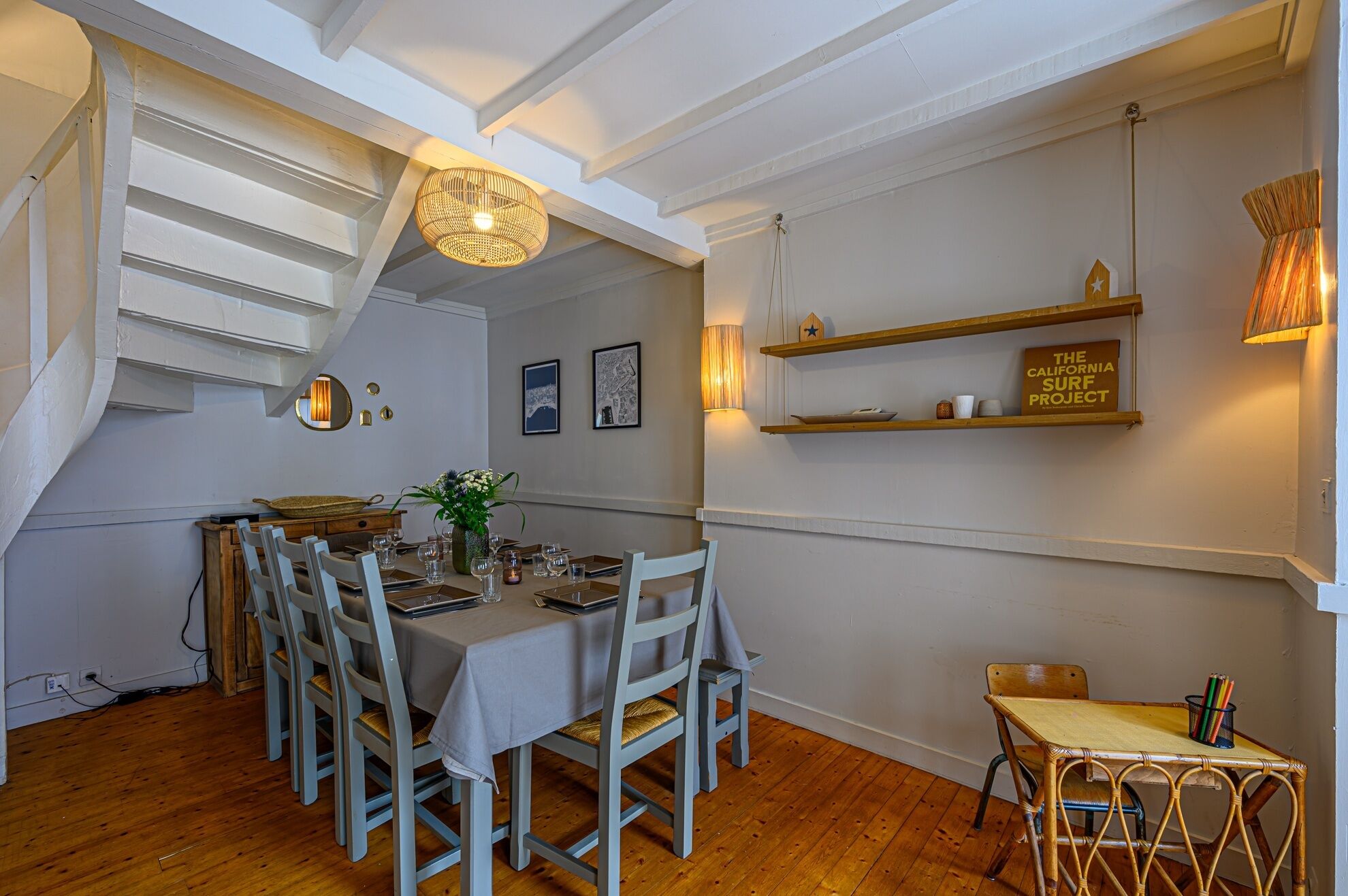 Un coin repas avec une longue table dressée pour six personnes, entourée de chaises. Des étagères murales présentent des objets décoratifs et un livre intitulé « The California Surf Project ».