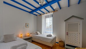 Un dormitorio con dos camas, piso de madera, una ventana con cortinas traslúcidas, un calendario colgado en la pared y una mesita con una silla.