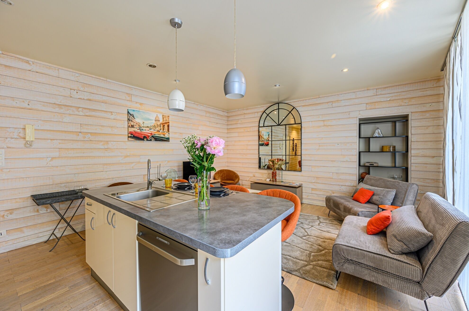 Een moderne keuken met een centraal eiland, een eetgedeelte met oranje stoelen, en een woonkamer met een grijze bank en oranje kussens.