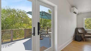 A room with a sliding glass door leading to an outdoor patio with a table and chairs, an air conditioning unit on the wall, and a window with a view of greenery.