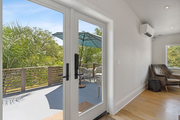 A room with a sliding glass door leading to an outdoor patio with a table and chairs, an air conditioning unit on the wall, and a window with a view of greenery. - Charming Studio Apartment C - Steps to Beach & Dining (St. Simons Island)