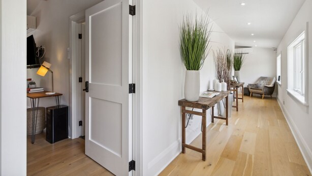 A hallway with a wooden floor, white walls, and a door leading to another room. There are potted plants and a small table with a lamp.
