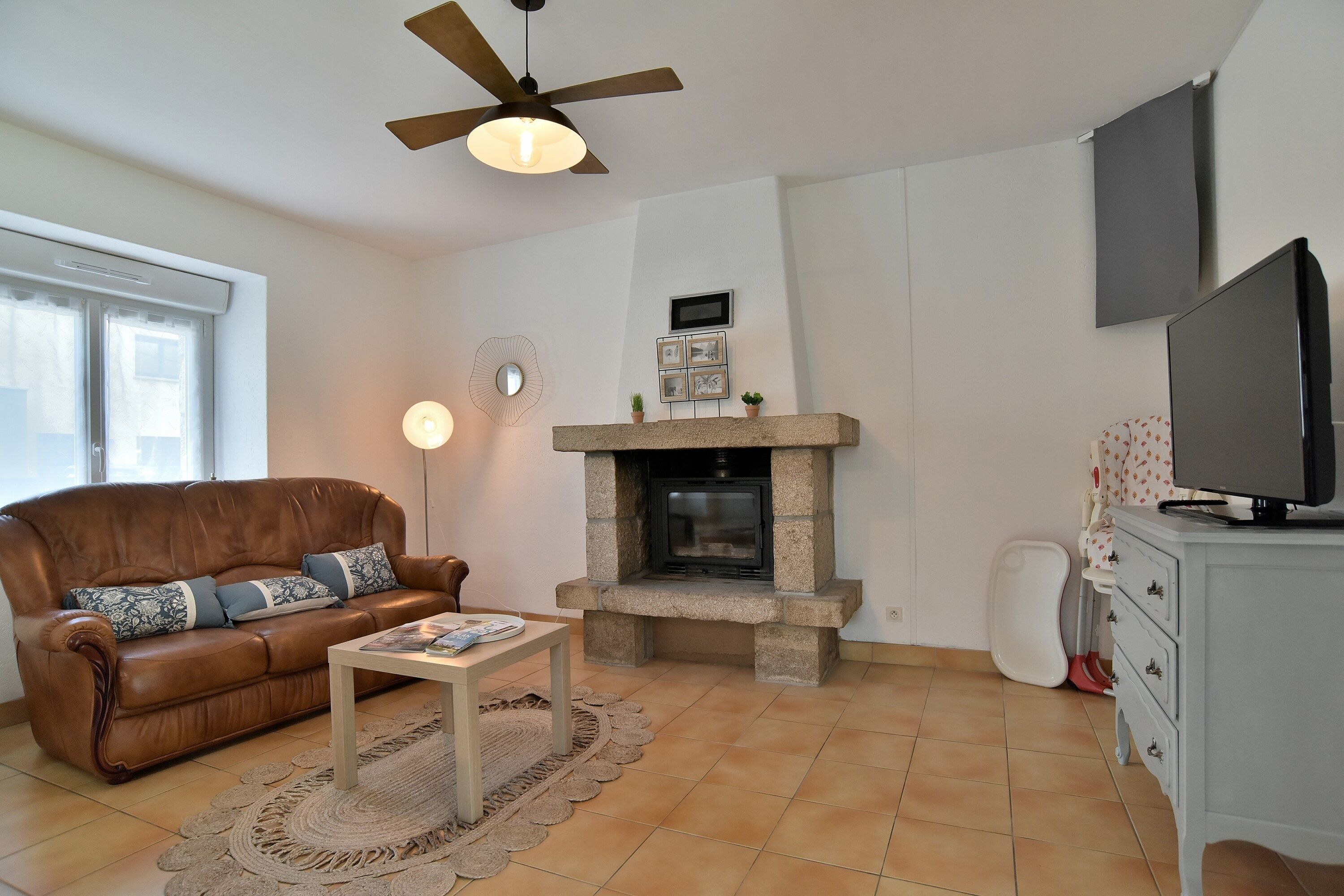 Un salon avec un canapé en cuir marron, une table basse, une cheminée en pierre, un ventilateur de plafond, un téléviseur posé sur un meuble et une armoire blanche.