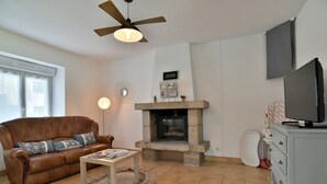 Ein Wohnzimmer mit braunem Ledersofa, einem Couchtisch, einem Kamin aus Stein, einem Deckenventilator, einem Fernseher auf einem Standfuß und einem weißen Schrank.