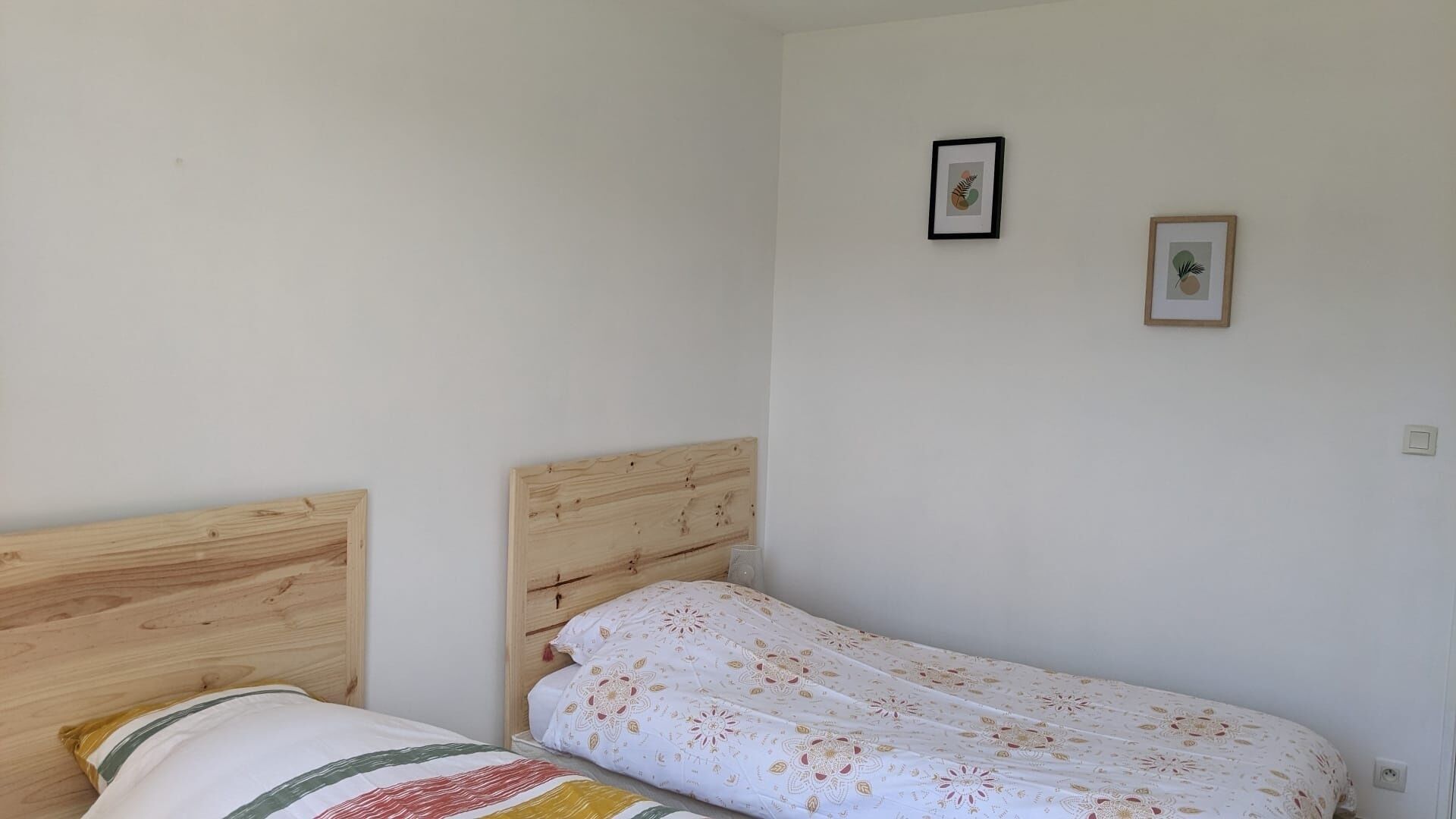 Een kleine kamer met een enkel bed, een houten hoofdbord, een wit sprei met bloemenprint en twee ingelijste foto's aan de muur.