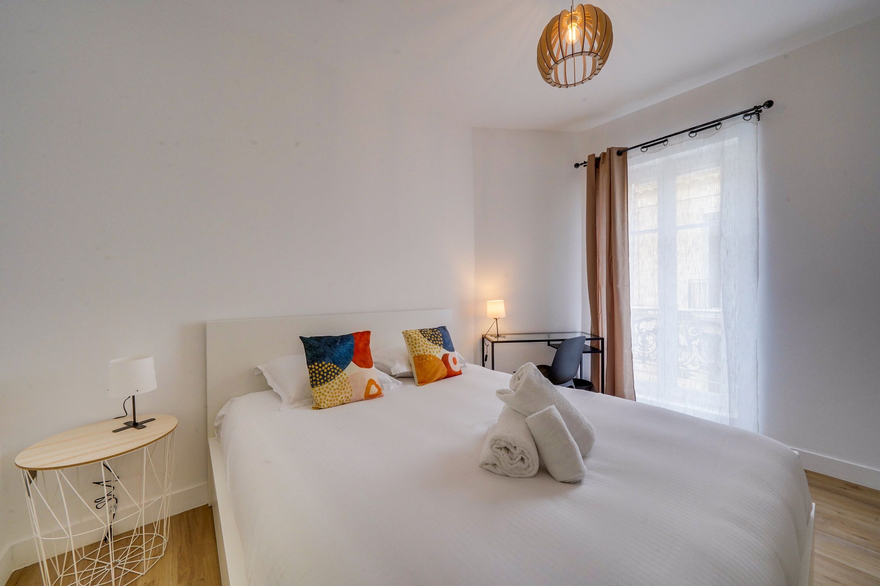 A hotel room with a large bed, a bedside table with a lamp, a desk with a chair, and a window with curtains.