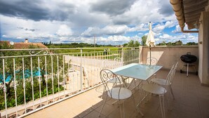 A balcony with a table and chairs, a grill, and a view of a swimming pool and greenery. - Côté Oliviers 6P - Appt avec piscine partagée (Beaucaire)