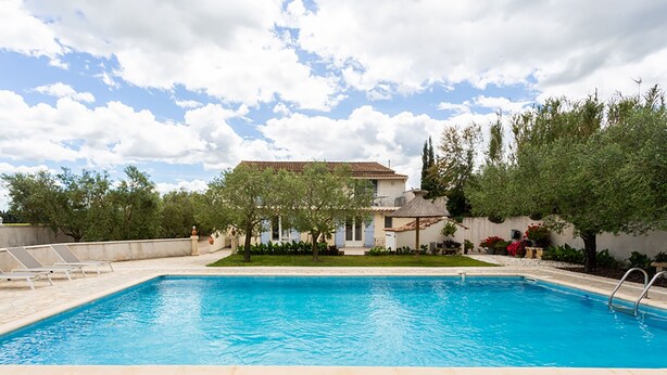 A swimming pool with a tiled deck, surrounded by greenery and a building with a tiled roof. - Côté Oliviers 6P - Appt avec piscine partagée (Beaucaire)