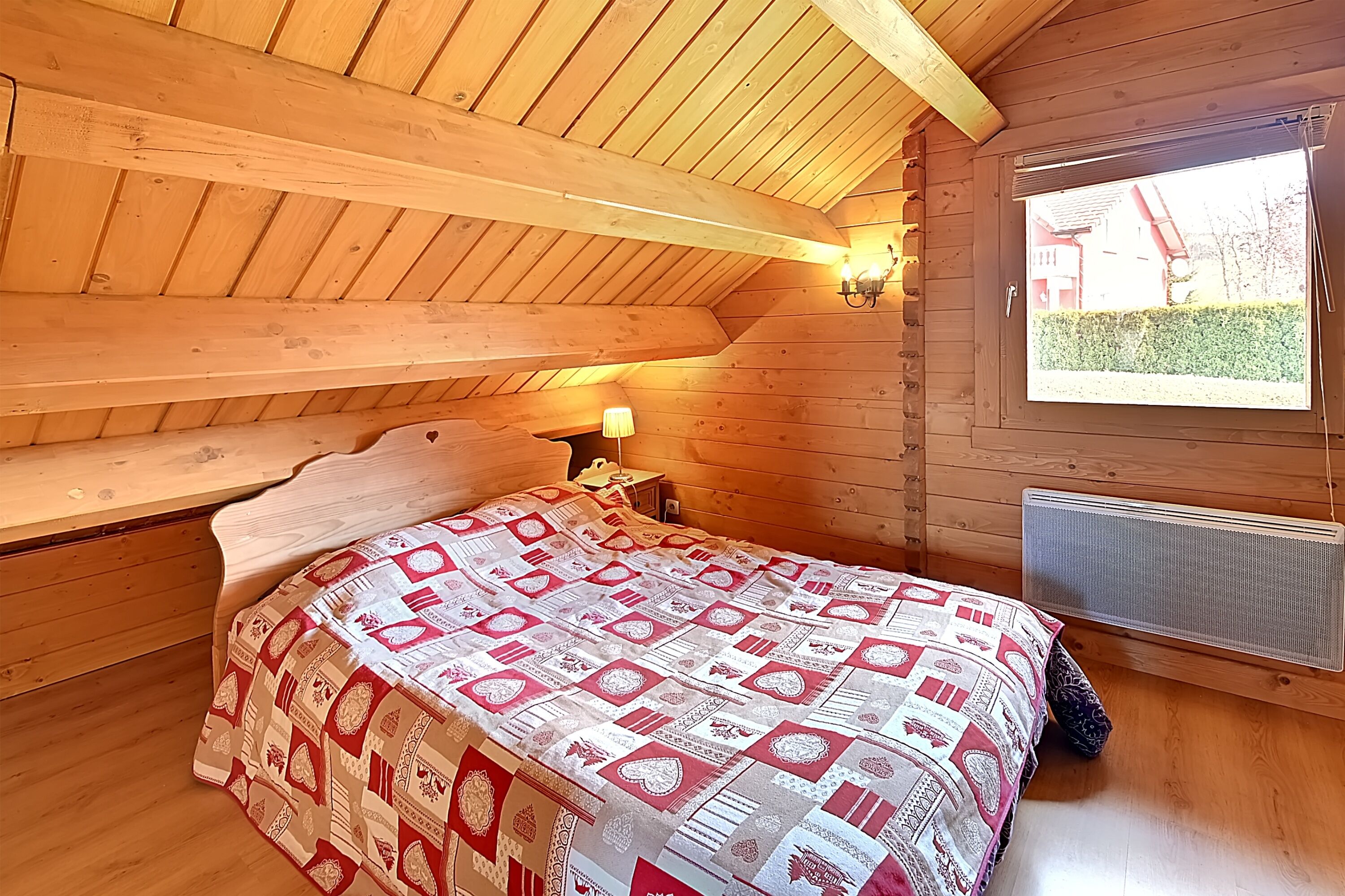A cozy wooden cabin bedroom with a bed, a nightstand, a lamp, and a window with a view of a house and hedge.