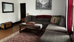 Una sala de estar con sofá, una mesa de centro, un tapete estampado, un cuadro en la pared y una mesita con lámpara.