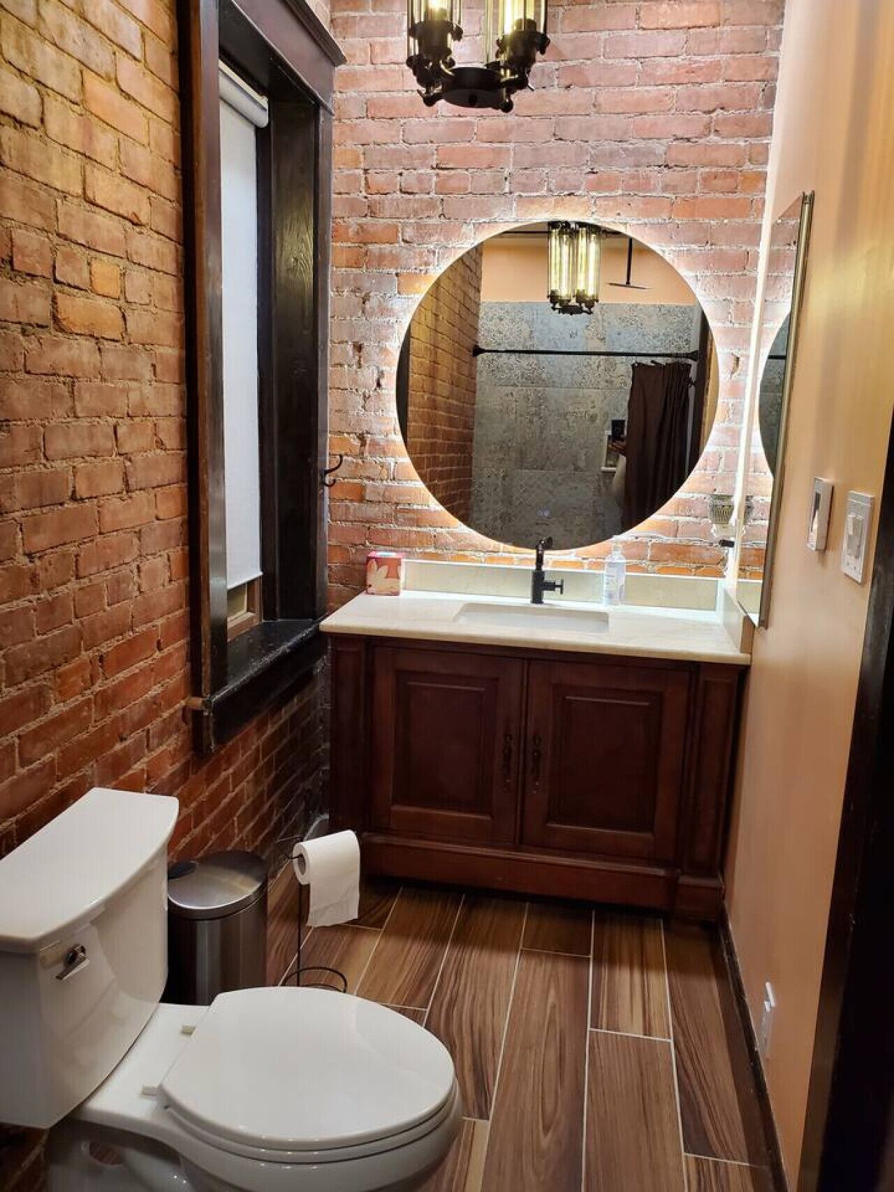 Un baño con pared de ladrillo, espejo redondo, lavamanos y retrete.