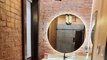 Un baño con pared de ladrillo, espejo redondo, lavamanos y retrete.