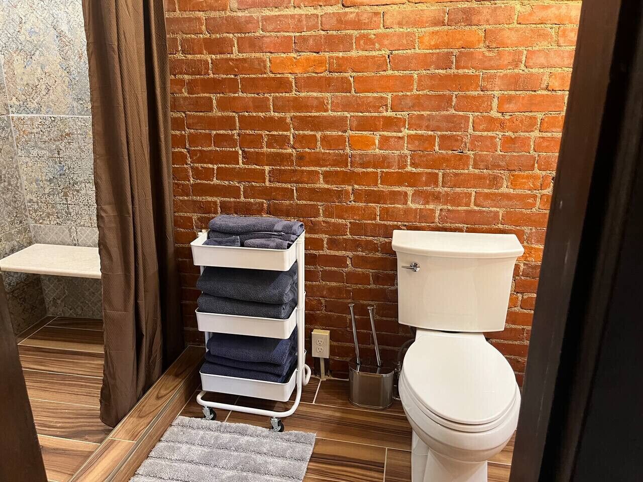 A bathroom with a brick wall, a white toilet, a towel rack with folded towels, and a wooden floor.