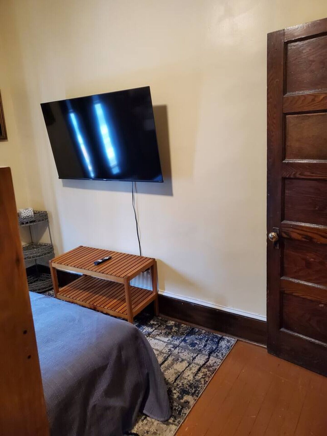 Una habitación con una cama, una pequeña mesa de madera, un televisor montado en la pared y una puerta de madera.