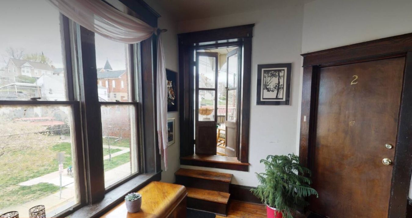 Una escalera conduce a un segundo piso con vistas a una zona residencial. Hay un banco al pie de la escalera, una planta en maceta a la derecha y una puerta con el número '2' también a la derecha.