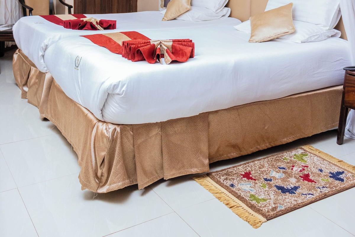 A large bed with white linens and a beige skirt, a wooden wardrobe, and a framed picture on the wall.