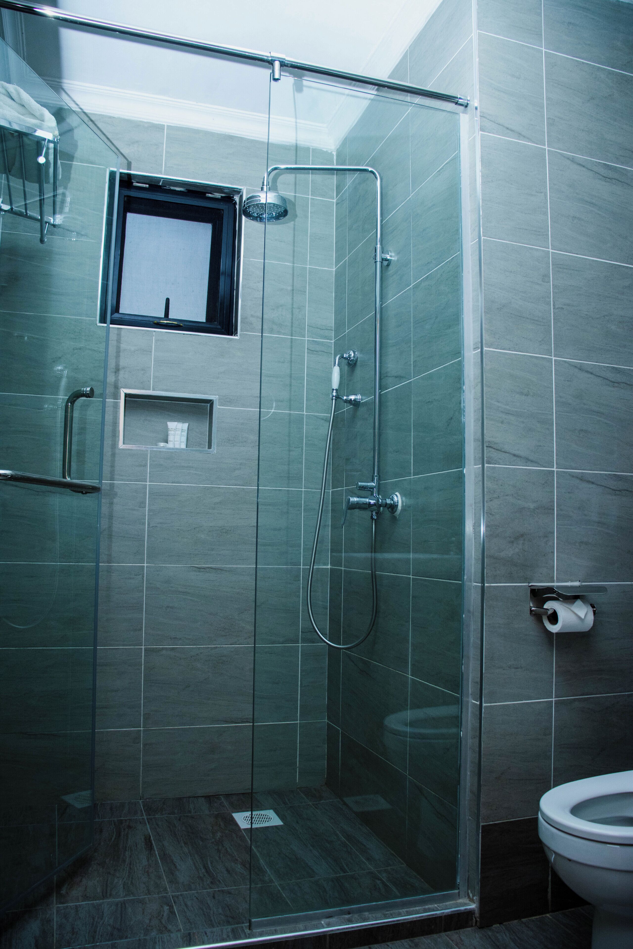 Ducha con paredes de vidrio, una ventana pequeña, un inodoro y un perchero para toallas.