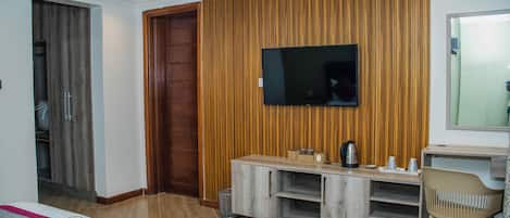 A hotel room with a bed, a TV mounted on a wooden panel wall, a small wooden side table, a chair, and a door leading to a bathroom.
