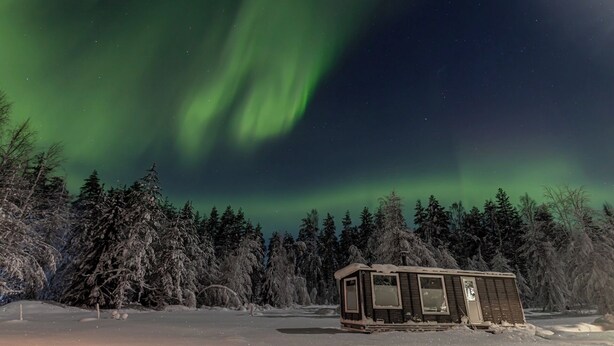 A small cabin in a snowy forest with a view of the Northern Lights.