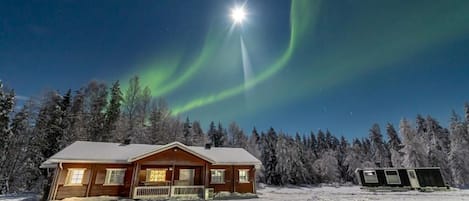 Une cabane en bois dotée d’un porche, entourée d’arbres couverts de neige et baignée par la clarté d’un ciel nocturne.