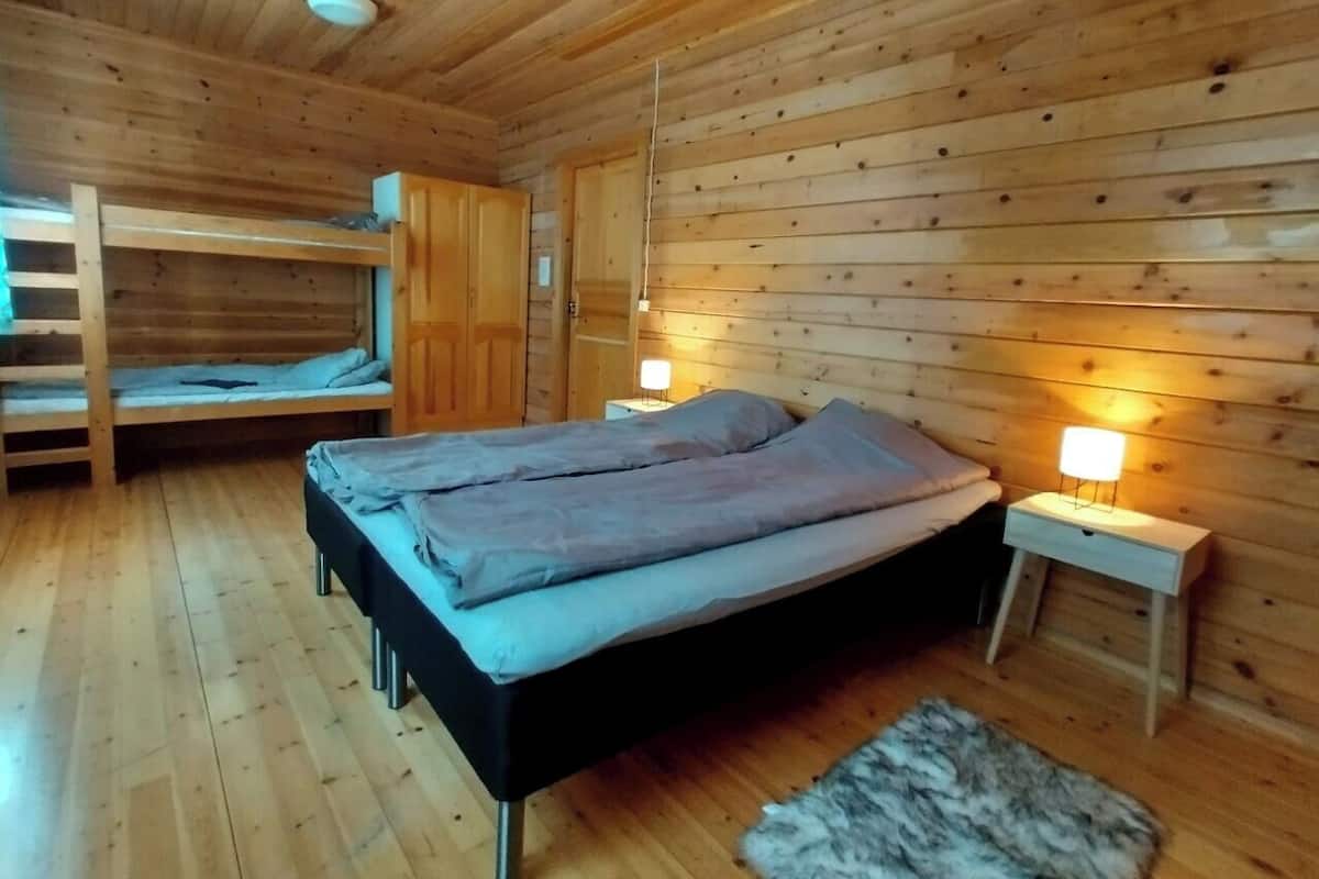 Habitació de cabana de fusta amb un llit superposat, un llit individual i una tauleta de nit amb una làmpada.