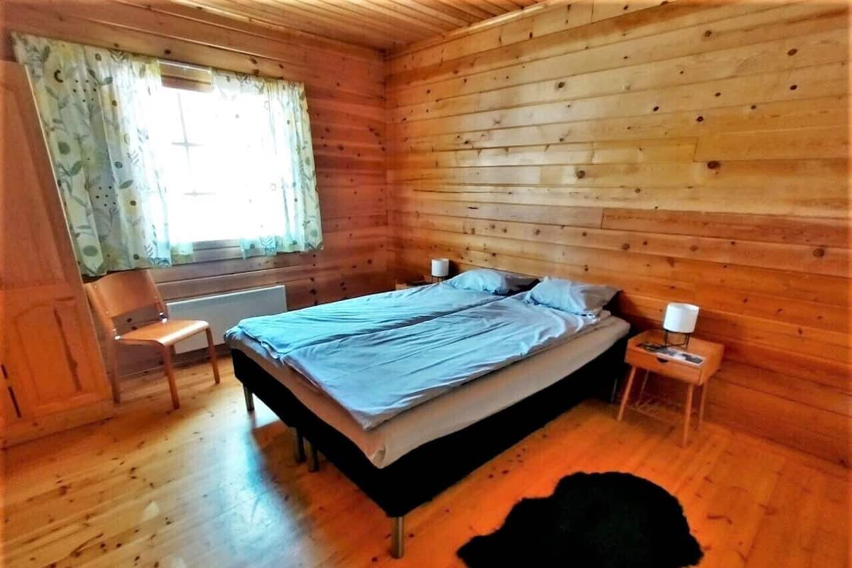 Habitació d'una cabana de fusta amb un llit, una cadira, una tauleta de nit i una finestra amb cortines.