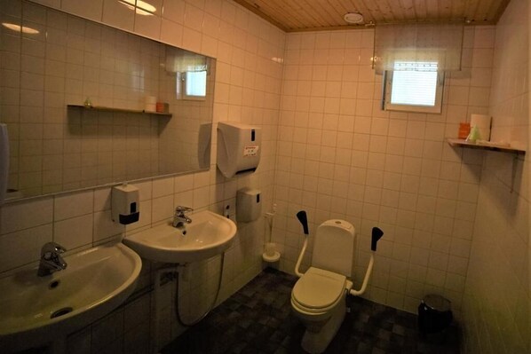 Un baño pequeño con lavamanos, inodoro y espejo. Hay repisas con artículos de aseo y una ventana con persianas.