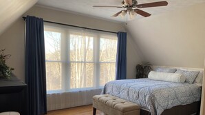 A bedroom with a large window, a ceiling fan, a bed with patterned bedding, a bench, and a radiator.