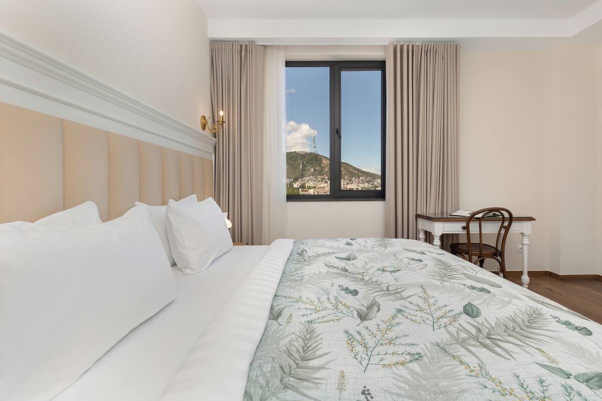 A hotel room with a large bed, a desk, and a window with a view of a cityscape and mountains.
