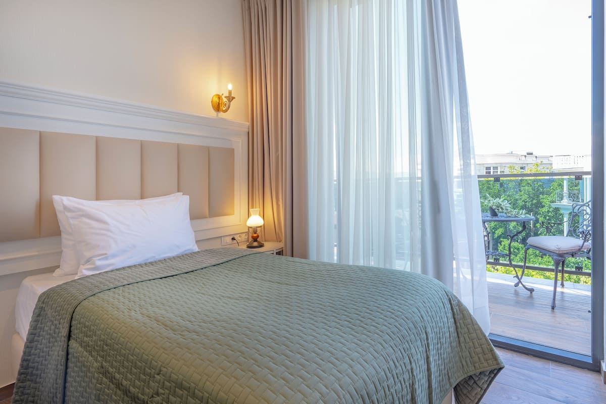 A hotel room with a bed, a nightstand with a lamp, and a view of a balcony with outdoor furniture.