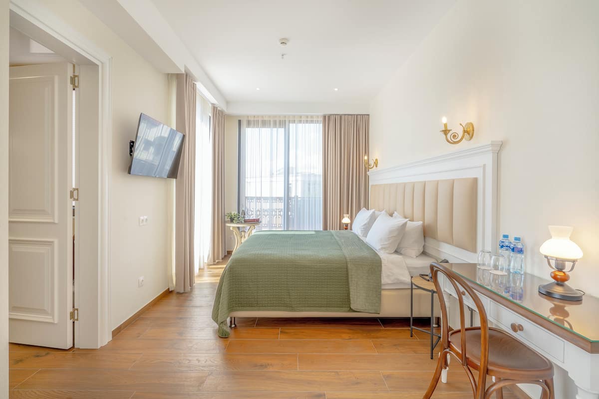 A hotel room with a bed, a desk with a chair, a television mounted on the wall, a fireplace, and a window with curtains.