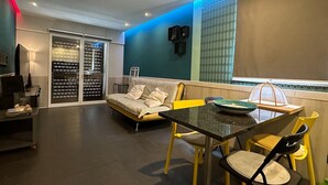 Sala de estar moderna com sofá, mesa de jantar e cadeiras amarelas. Há uma TV e uma janela com persianas.