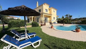 A poolside area with lounge chairs, a thatched umbrella, and a house with a tiled roof. - Villa Sao Miguel (Albufeira)