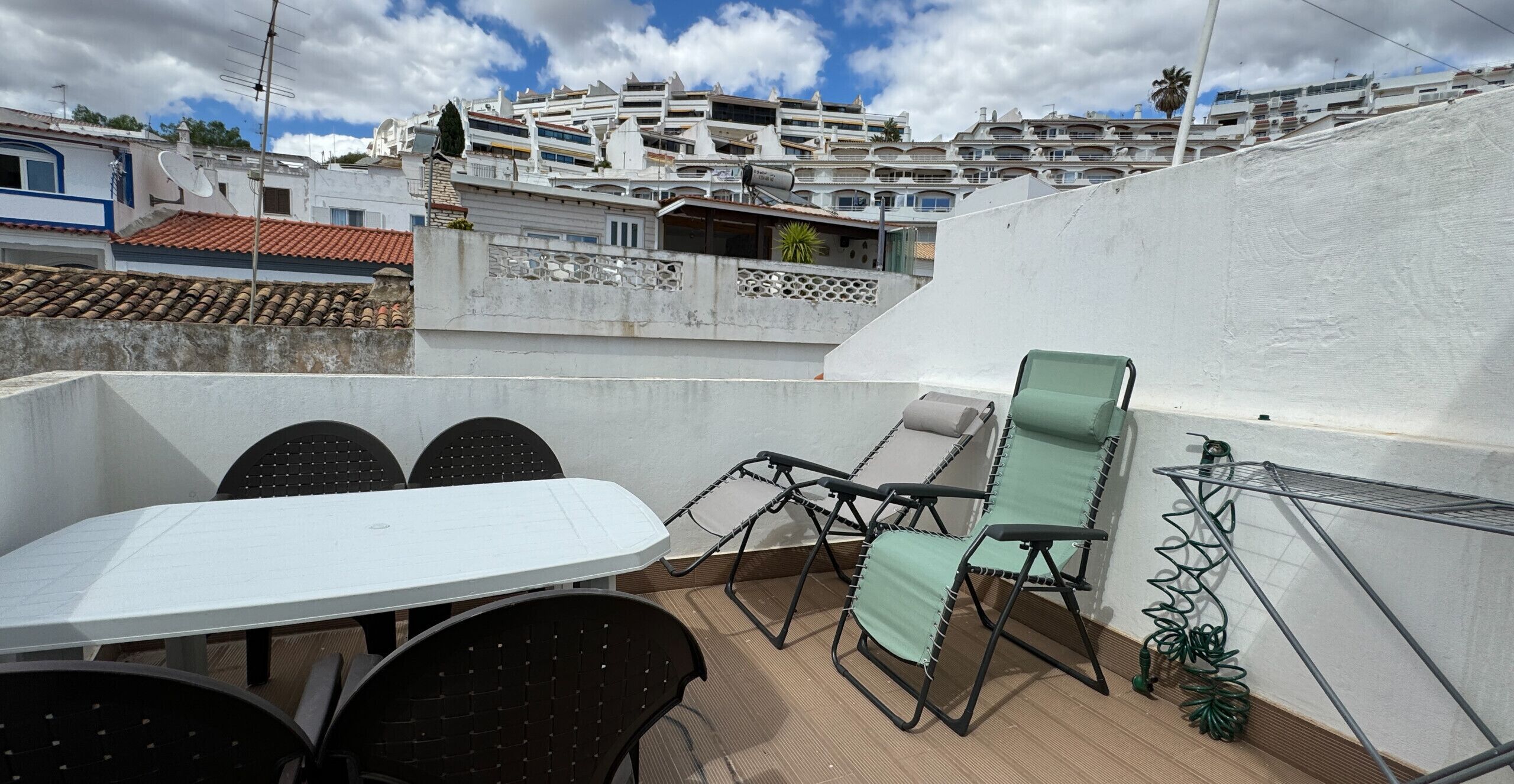Eine Dachterrasse mit Tisch und Stühlen, einem Metallregal und Blick auf ein Wohngebiet mit weißen Gebäuden.