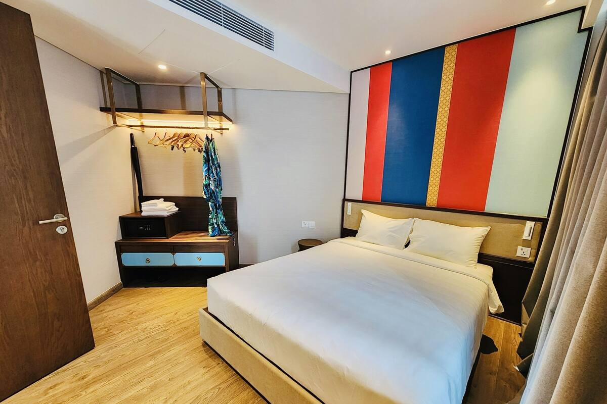 Une chambre d’hôtel moderne avec un grand lit, une commode en bois, un miroir et un panneau mural aux couleurs vives.