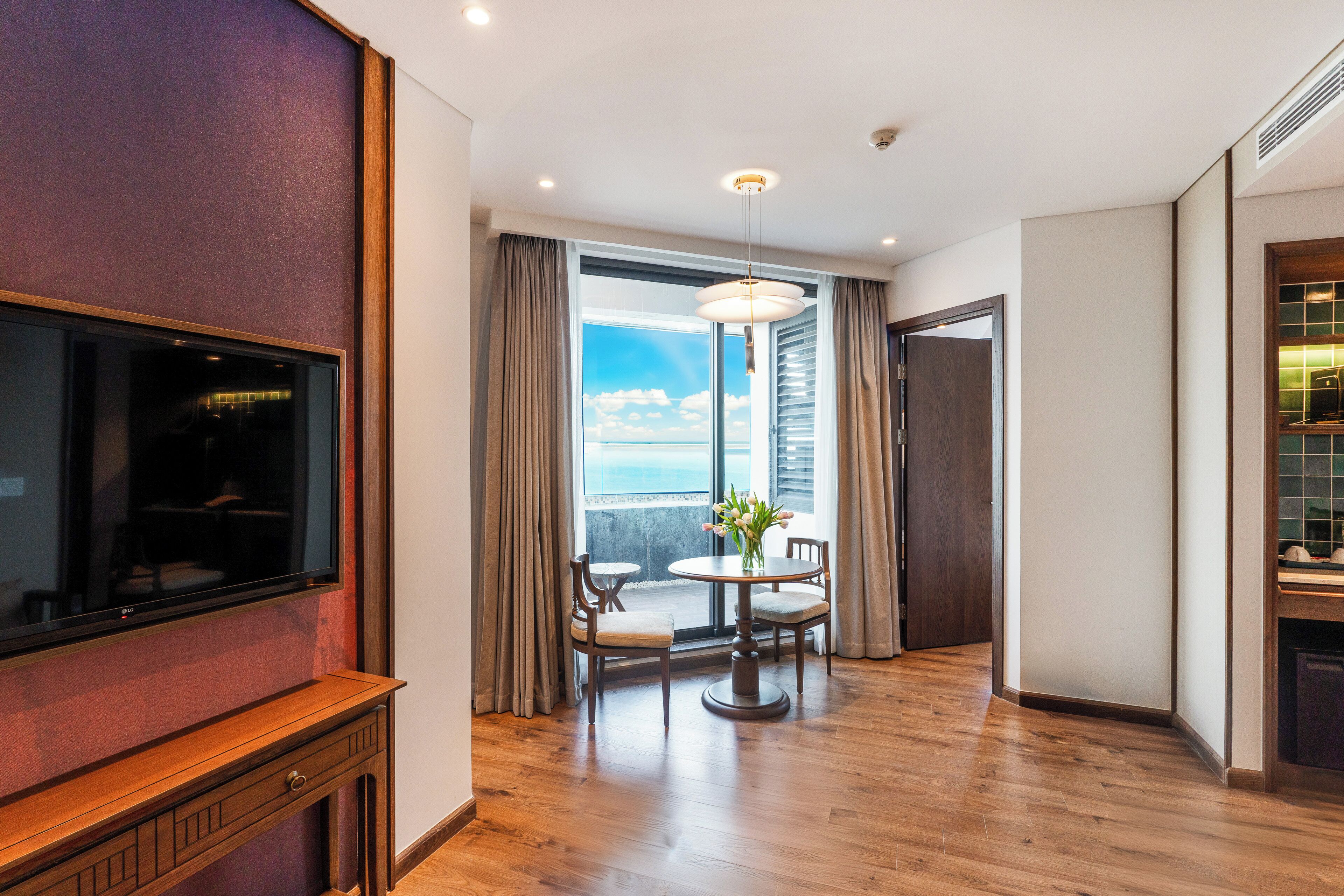 A hotel room with a flat-screen TV, a dining table with chairs, a large window with curtains, and a ceiling light.