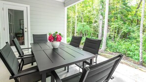 En overdækket terrasse med bord og stole, en blomstervase og udsigt til et skovområde.