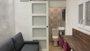 A compact room with a grey sofa, a wooden table, and a red chair. A sliding door leads to a bathroom with a toilet and sink.