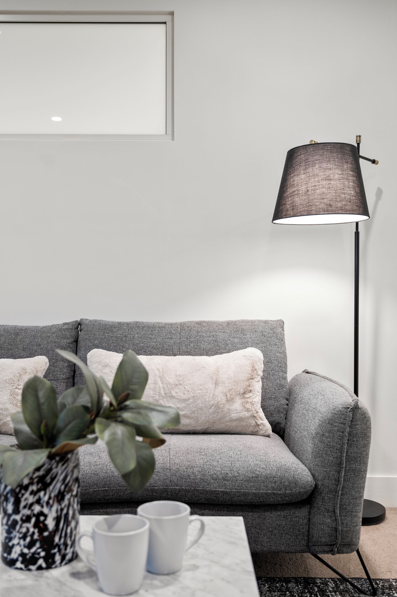 Ein modernes Wohnzimmer mit einem grauen Sofa, einer Stehlampe, einer Topfpflanze und zwei weißen Tassen auf einem Marmortisch.
