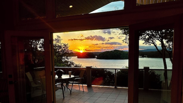 Una sala de estar con sofá y mesa, vista a un atardecer sobre el agua, y un balcón con mesa y sillas.