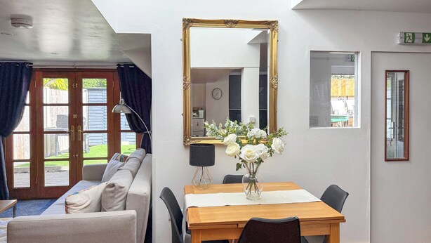 A room with a sofa, a dining table with chairs, a mirror, and a vase with flowers. - 2 bedroom accommodation in Carterton (Carterton)