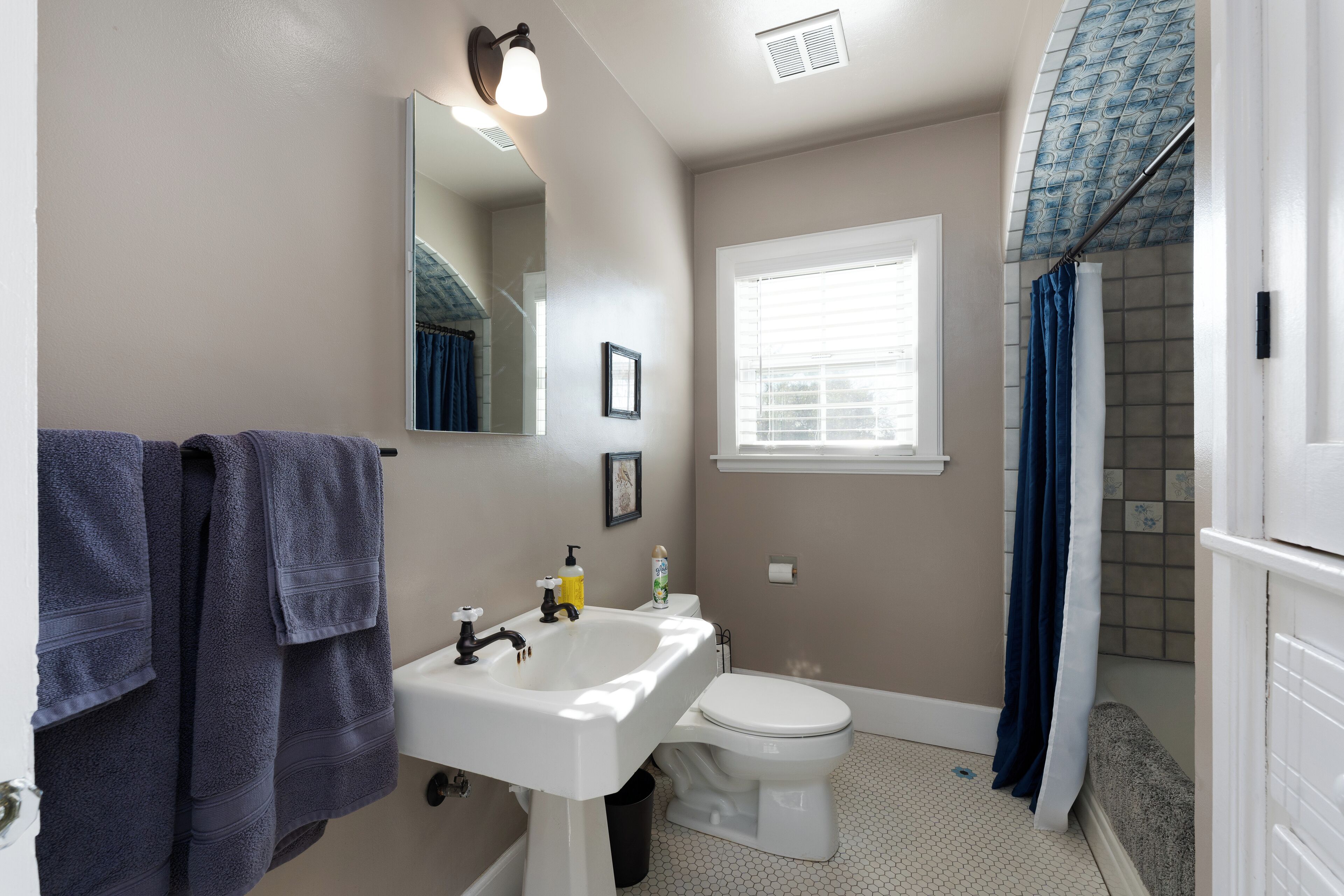 A bathroom with a white sink, toilet, and shower. There is a window with blinds, a mirror, and a towel rack with towels.