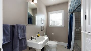 A bathroom with a white sink, toilet, and shower. There is a window with blinds, a mirror, and a towel rack with towels.