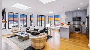 Una sala moderna con un sofá, sillones y una mesa de centro. Cuenta con una cocina equipada con microondas, estufa y fregadero. Amplias ventanas ofrecen vistas al atardecer.
