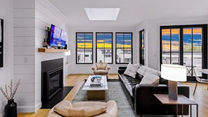 Una sala de estar moderna con chimenea, un sofá, una mesa de centro y un televisor. Amplias ventanas con vistas al cielo y edificios.