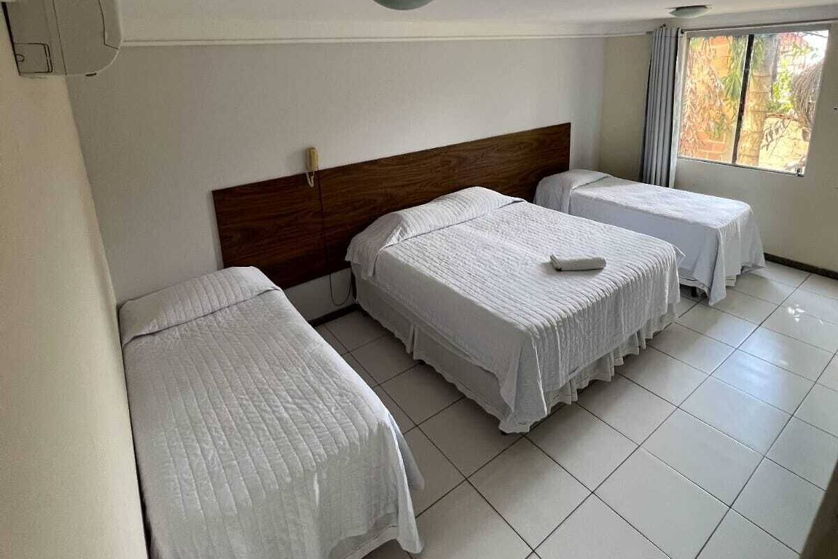 Una habitación de hotel con tres camas individuales, un cabecero de madera, un ventanal con cortinas y suelo de baldosas.